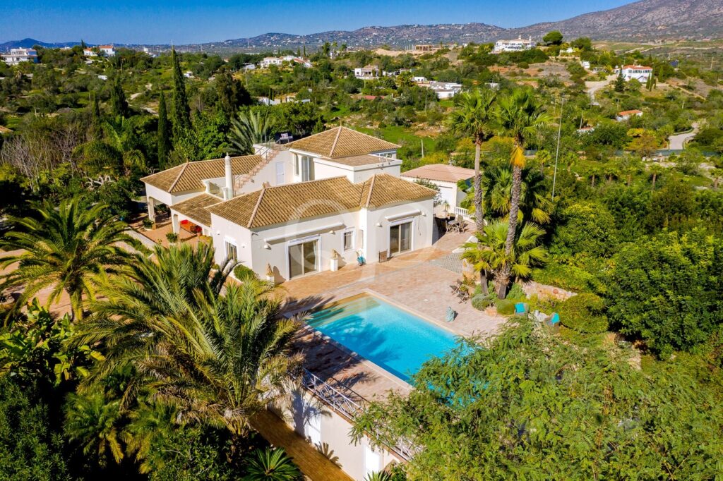 Villa méditerranéenne blanche aux toits en terre cuite, agrémentée de palmiers luxuriants et dotée d'une piscine bleue rectangulaire, située sur une colline ensoleillée.