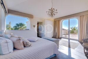 Bright bedroom with a white quilted bed, pastel pillows, and a chandelier, open arched windows showing a sunny outdoor scene.