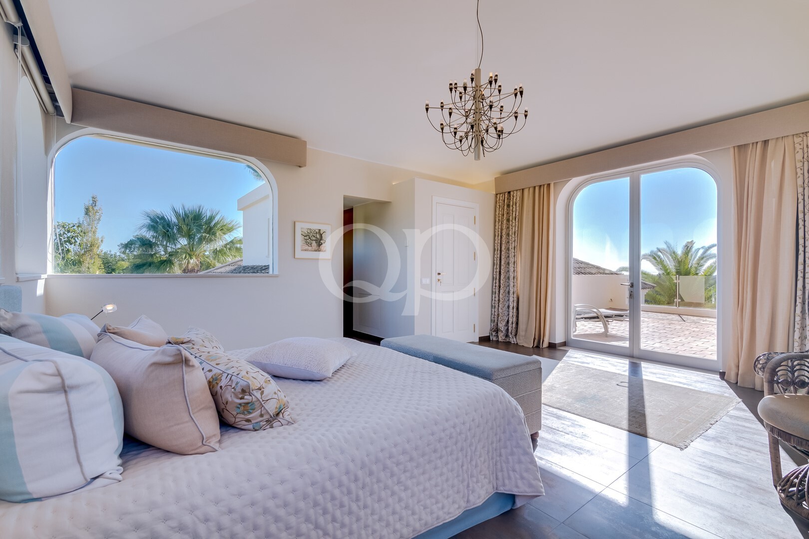 Bright bedroom with a white quilted bed, pastel pillows, and a chandelier, open arched windows showing a sunny outdoor scene.