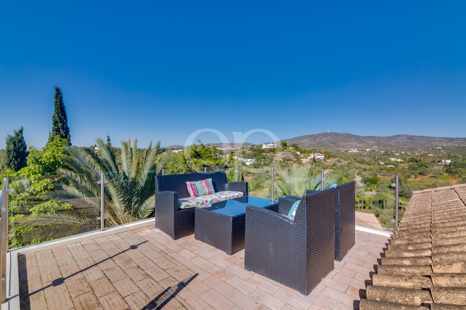 Sunny rooftop patio with black wicker furniture and colorful cushions, glass railing, and a rolling hillside view beneath a clear blue sky.