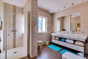 Modern beige-toned bathroom with glass shower, double sinks, and open towel storage under the countertop