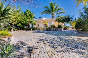 Stone-paved circular driveway leading to a cream-colored villa with tiled roof and palm trees on a sunny day.