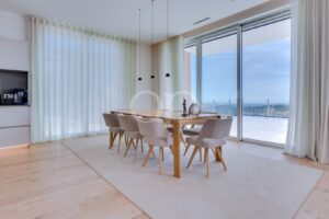 Bright dining room with a wooden table and eight beige upholstered chairs, large windows, sheer curtains, and an ocean view outside.