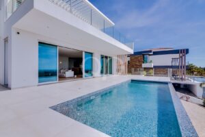 Contemporary white villa with a long swimming pool, glass balcony rails, and sliding glass doors opening to an interior living area.