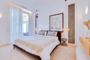 Modern white bedroom with a low dark bed, beige duvet, two patterned throw pillows, and pendant lamps on either side.