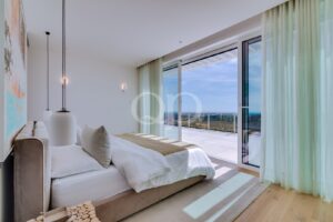 Bedroom with a low, upholstered bed facing glass doors that open to a balcony with a panoramic coastal view and mint curtains