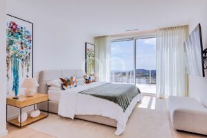 Bright modern bedroom with a beige upholstered bed, white linens, a green throw, and colorful abstract paintings flanking the bed on a side table with a lamp nearby.