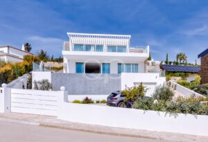 Modern white multi‑level house with glass balconies and a blue sky backdrop; a gated front yard and a parked SUV visible.