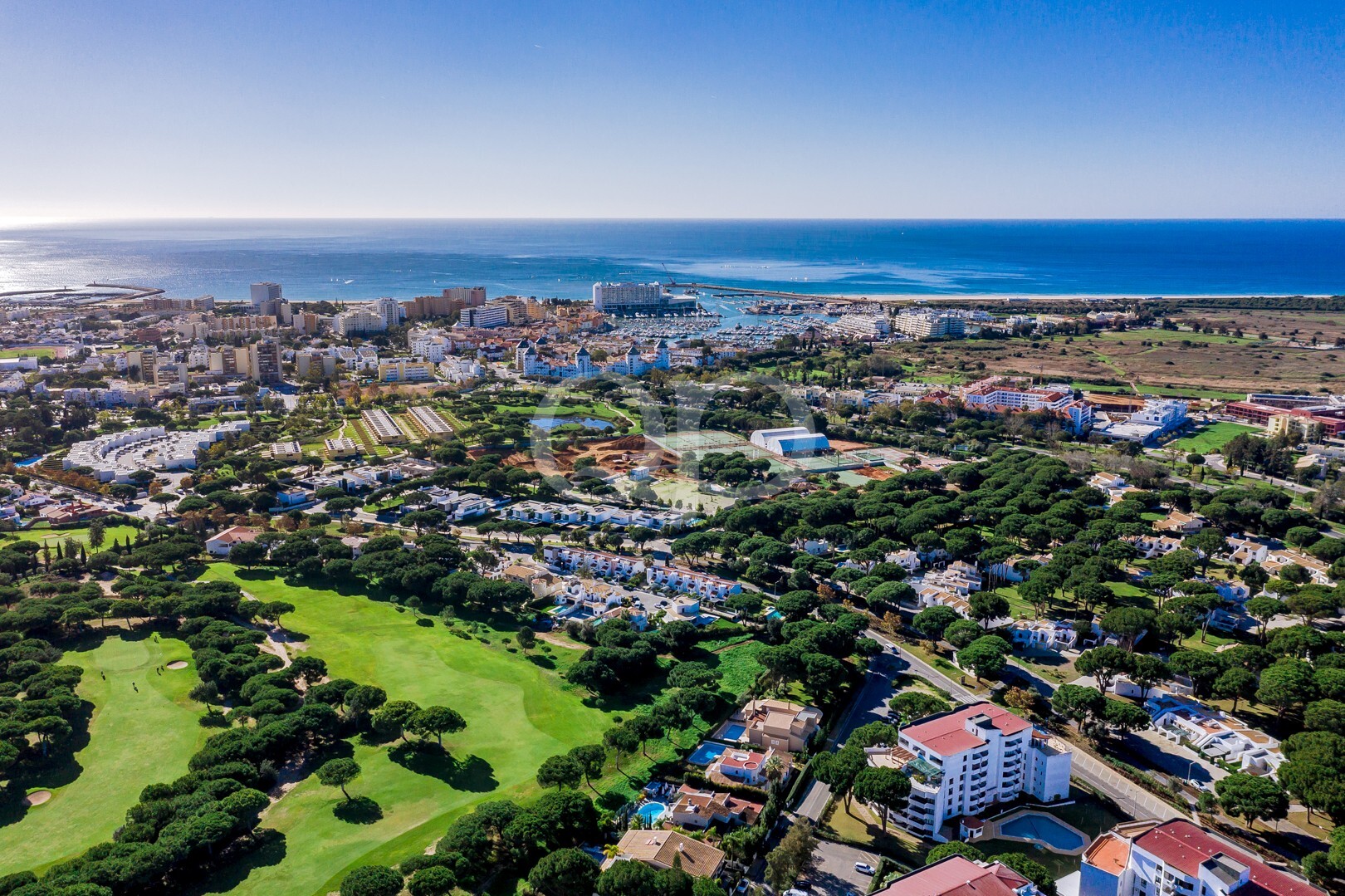 Luftaufnahme einer Küstenstadt mit einem großen, grünen Golfplatz im Vordergrund, Wohnblocks und einem Hafen am blauen Meer.
