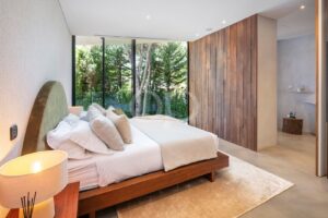 Modernes Schlafzimmer mit einem Podestbett, weißer Bettwäsche und beigen Kissen, mit Blick auf eine große Glaswand, durch die man den Garten sehen kann.