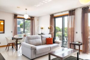 Bright modern living room with a gray sofa, orange throw pillow, and glass doors opening to a patio. The space includes a dining table with white chairs and sheer brown curtains.