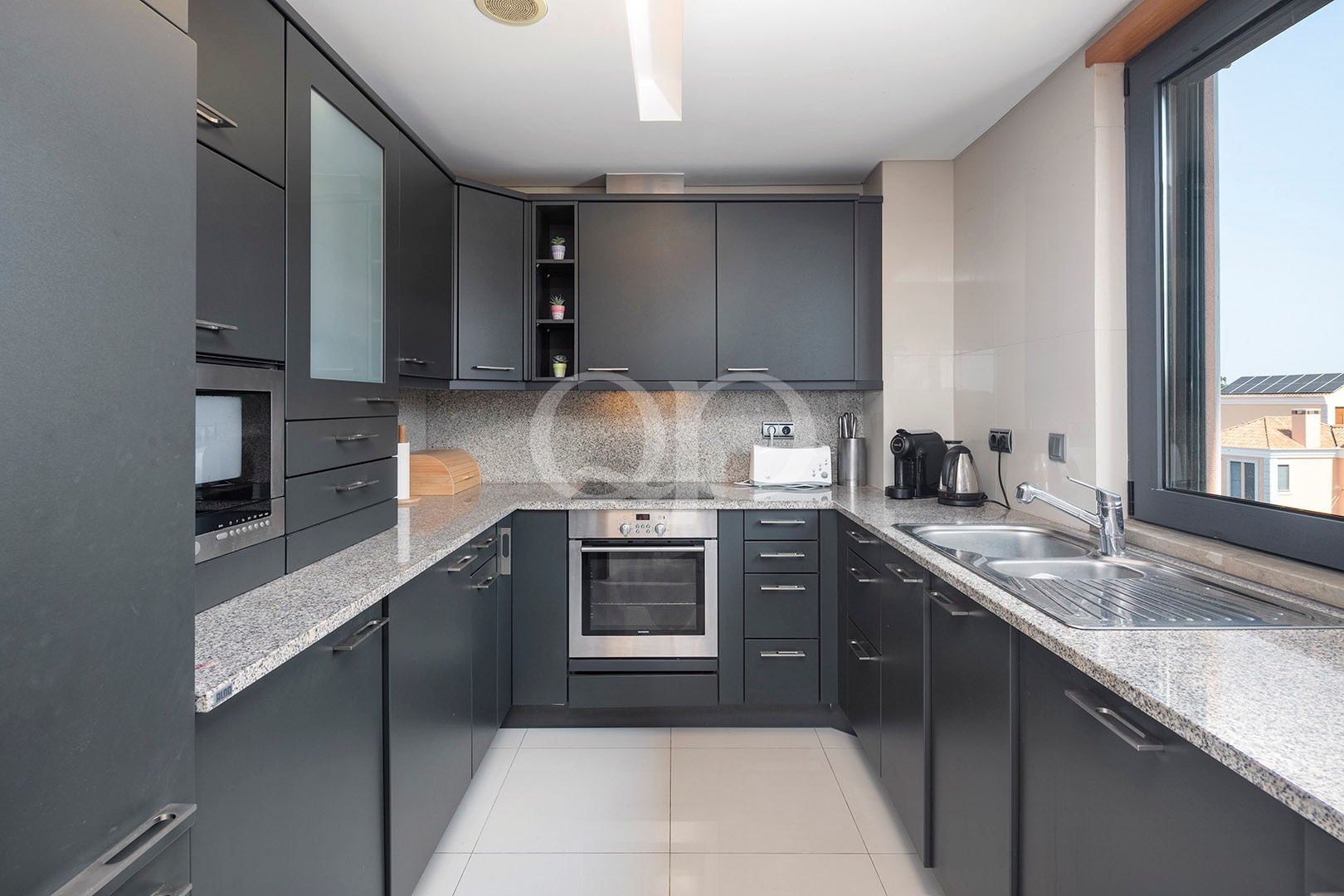 Modern L-shaped kitchen with dark grey cabinets, granite countertops, built-in oven, microwave, and a double sink by a large window.