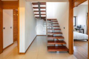 Open hallway with a floating wooden staircase at center, white walls and wood-framed doorways.