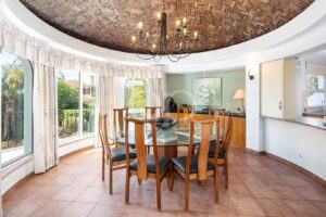 Round glass dining table with eight wooden chairs under a black chandelier in a sunlit dining area with a curved brick ceiling and large windows.