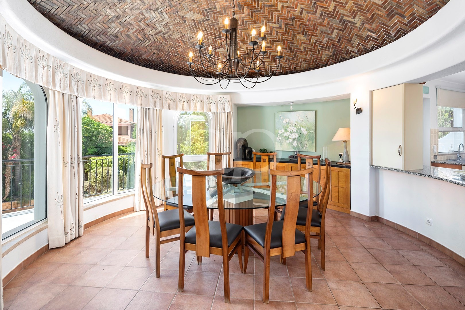 Round glass dining table with eight wooden chairs under a black chandelier in a sunlit dining area with a curved brick ceiling and large windows.