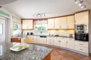 Bright L-shaped kitchen with cream cabinets, granite counters, and stainless appliances under track lighting, island in foreground.