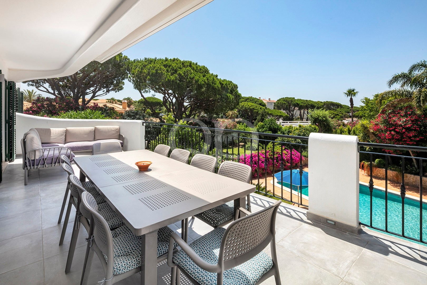Outdoor balcony with a long dining table and multiple chairs, overlooking a pool and lush garden under a clear blue sky.