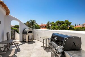Sunny rooftop patio with a pizza oven, gas grill, and beige seating under a clear blue sky.