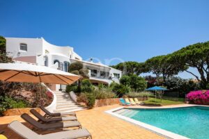 White Mediterranean-style villa with a pool, lounge chairs, and umbrellas under a clear blue sky.