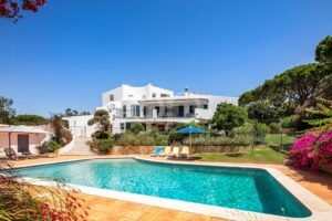 White villa with curved balconies overlooking a bright pool, blue sky above, and lounge chairs under a blue umbrella.