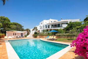 White villa with curved balconies and a clear blue sky, overlooking a turquoise pool with lounge chairs and umbrellas.