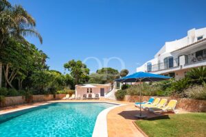 Sunlit hotel pool area with lounge chairs, a blue umbrella, and a white Mediterranean-style building along a stone wall garden boundary.