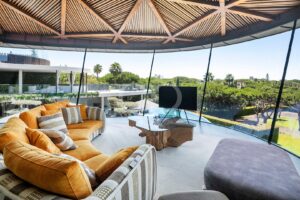 Ein luxuriöses, geschwungenes orangefarbenes Sofa mit gemusterten Kissen in einem sonnendurchfluteten Wohnzimmer mit Glaswänden und Blick auf einen grünen Garten und den Poolbereich im Freien.