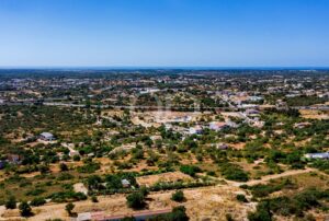 Aerial view of a semi-arid town with scattered houses, roads, and sparse trees, extending toward a distant coastline under a clear blue sky.