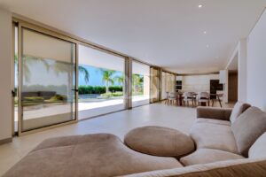 Bright, open-plan living space with large floor-to-ceiling sliding glass doors opening to a sunny outdoor patio and pool area; a beige sectional sofa is in the foreground.