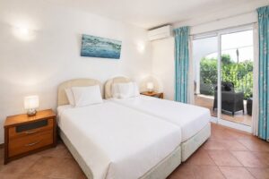 Twin-bed hotel room with white linens, beige headboards, and matching nightstands on either side, plus a sliding door to a balcony.