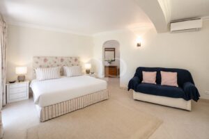 A bright hotel bedroom with a large bed, floral headboard, striped pillows, and two white nightstands with lamps on either side.