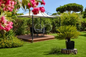 Essbereich im Garten unter einem Pavillon mit weißem Baldachin auf einer Holzterrasse, mit Tisch und Stühlen, umgeben von üppigem Grün und rosa Blumen.