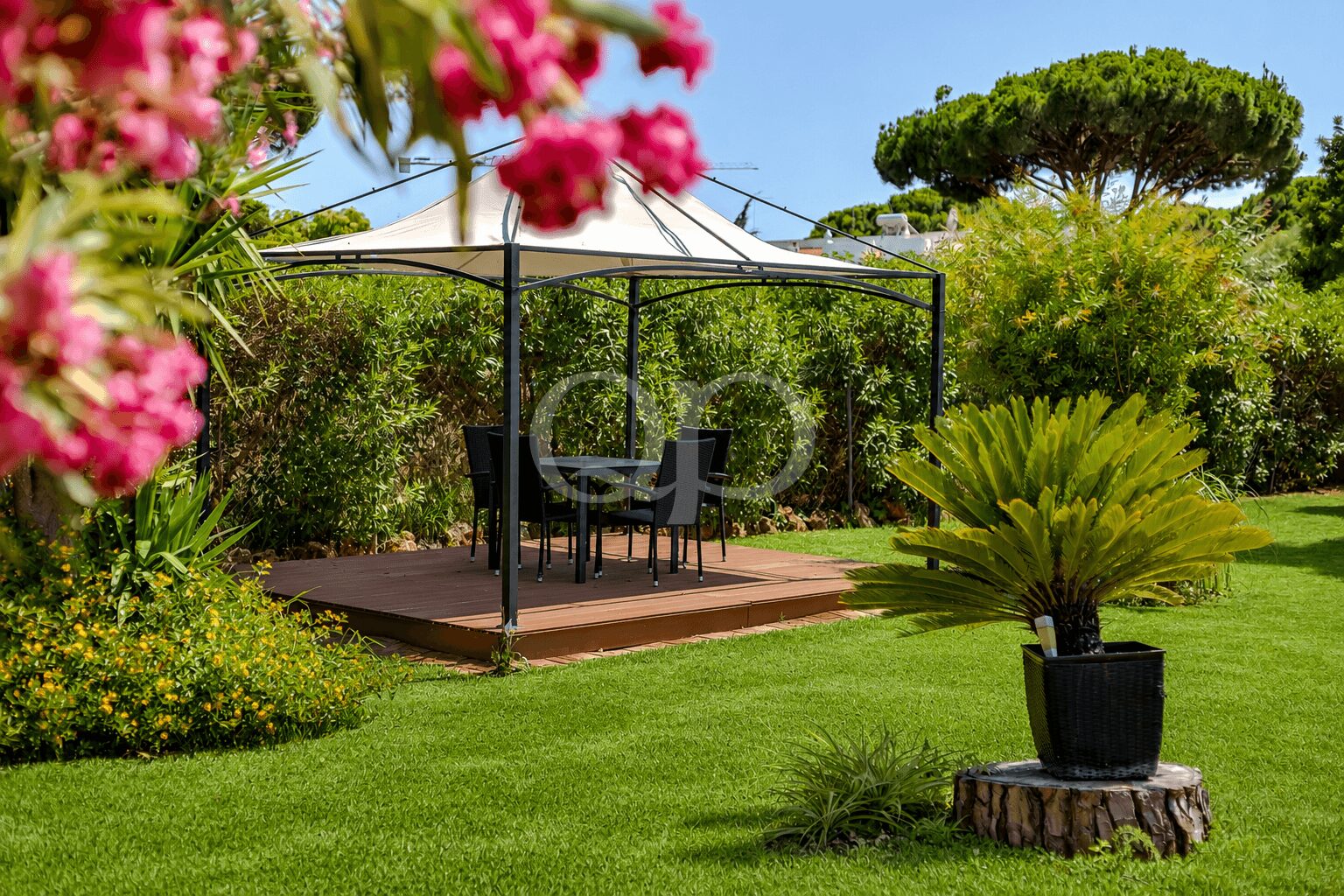 Essbereich im Garten unter einem Pavillon mit weißem Baldachin auf einer Holzterrasse, mit Tisch und Stühlen, umgeben von üppigem Grün und rosa Blumen.