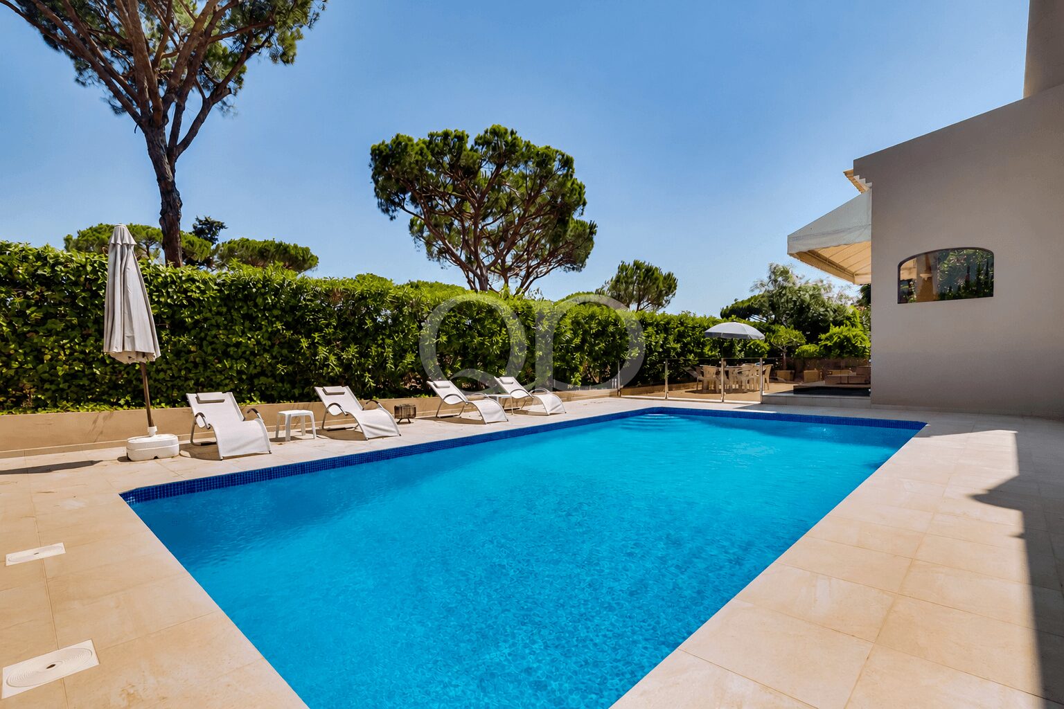 Ein rechteckiger blauer Pool mit beigem Umrandungsbereich und weißen Liegestühlen entlang einer grünen Hecke unter strahlend blauem Himmel.