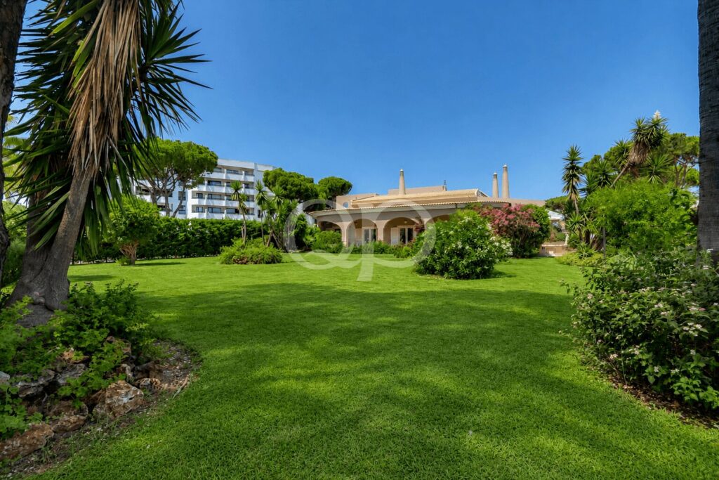 Ein üppiger Resortgarten mit einer Rasenfläche, Palmen und einer beigen Villa mit Bogenfenstern im Hintergrund unter einem strahlend blauen Himmel.