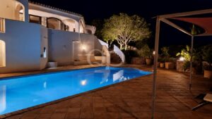Night-time view of a blue-lit rectangular pool beside a white Mediterranean villa with arches and stairs, surrounded by plants and warm outdoor lighting.