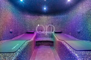 Colorful tiled steam room with two massage beds and a central bench, illuminated by blue and purple lights at nightfall-like ambience.