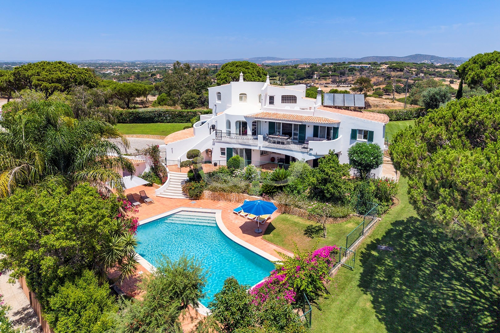 Aerial view of a white Mediterranean villa with terracotta roof, curved balconies, and a turquoise pool surrounded by lush gardens.