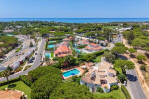 Aerial view of a coastal residential area with red-tiled-roof houses, large pools, and lush trees, leading to the blue sea on the horizon.
