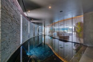 Ein von gebogenem Glas umschlossener Poolbereich mit einer dunklen Steinmauer auf der linken Seite und einem dahinter liegenden Wohnzimmer.