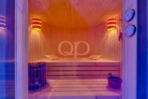 Innenansicht einer Holzsauna mit zweistufigen Bänken, einem Ofen auf der linken Seite und einem Eimer auf dem Boden unter warmer Beleuchtung.