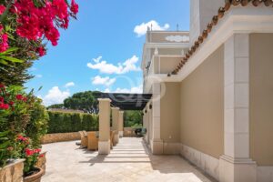 Terrasse d'une villa beige à colonnes, avec une pergola sombre et un coin salon extérieur sous un ciel bleu et ensoleillé, avec des fleurs roses à gauche.