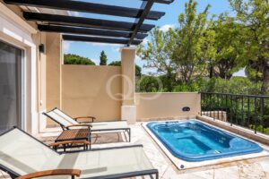 Une terrasse ensoleillée avec un petit jacuzzi bleu, des murs beiges et deux chaises longues au plateau en verre, sous une pergola noire, donnant sur la verdure.