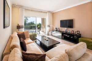 Salon lumineux et moderne, avec un canapé modulable beige, une table basse en verre, une télévision fixée au mur sur un mur d'accent couleur beige clair, ainsi qu'une porte-fenêtre.