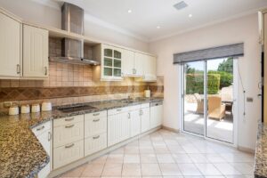 Cuisine lumineuse avec des meubles de cuisine couleur crème, des plans de travail en granit et une crédence carrelée, ainsi qu'une hotte en inox au-dessus de la cuisinière et une porte coulissante donnant sur une terrasse extérieure équipée de chaises en osier.