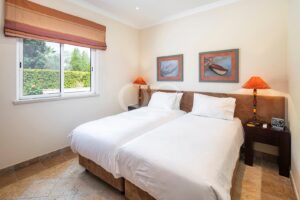 Deux lits jumeaux avec une parure de lit blanche et une tête de lit commune marron, dans une chambre d'hôtel lumineuse équipée de deux tables de chevet et de lampes orange.