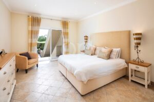 Chambre d'hôtel lumineuse avec un grand lit beige, des oreillers rayés et des tables de chevet assorties, flanquées de lampes décoratives de chaque côté de la tête de lit, ainsi qu'une porte-fenêtre donnant sur le balcon.