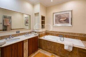Salle de bains carrelée de beige, avec un meuble-lavabo en marbre à double vasque, un grand miroir et une baignoire encastrée le long du mur carrelé.