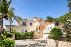 Peach-colored Mediterranean villa with tiled roof, arched entrance, stone retaining wall, and palm trees against a bright blue sky.