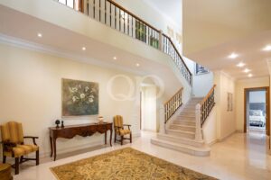 Elegant two‑story foyer with curved marble staircase, cream walls, and a wooden console table flanked by striped chairs.